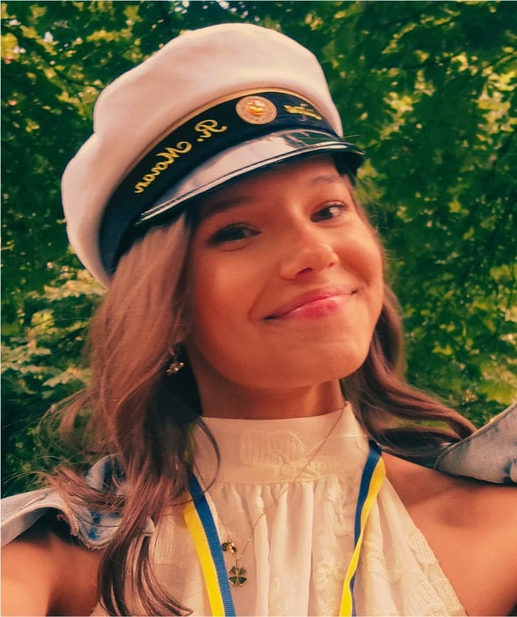 Ciara smiling in a white Swedish graduation cap, sunlit green trees behind her