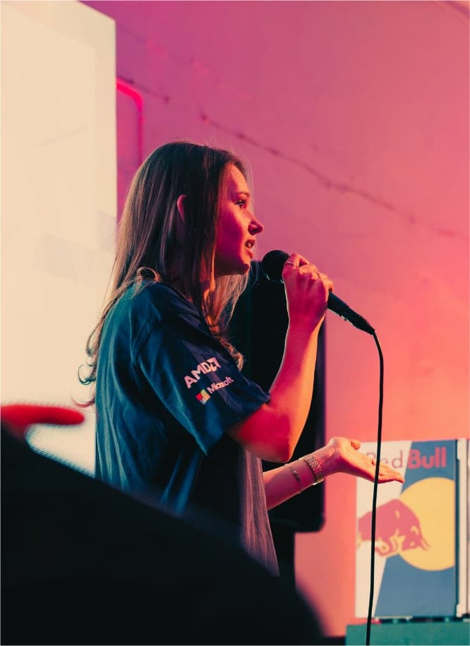 Ciara speaking into a microphone on stage, lit pink and red at a hackathon